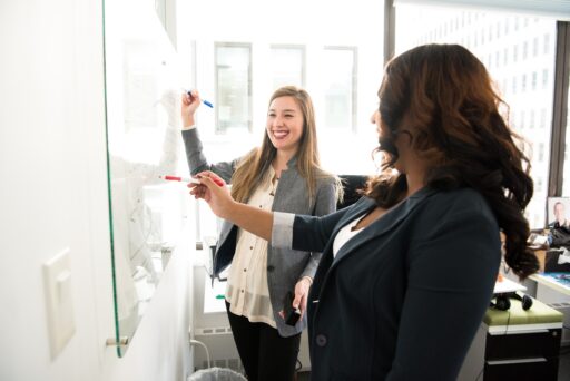 Two people stand next to a white board writing and pointing to it.